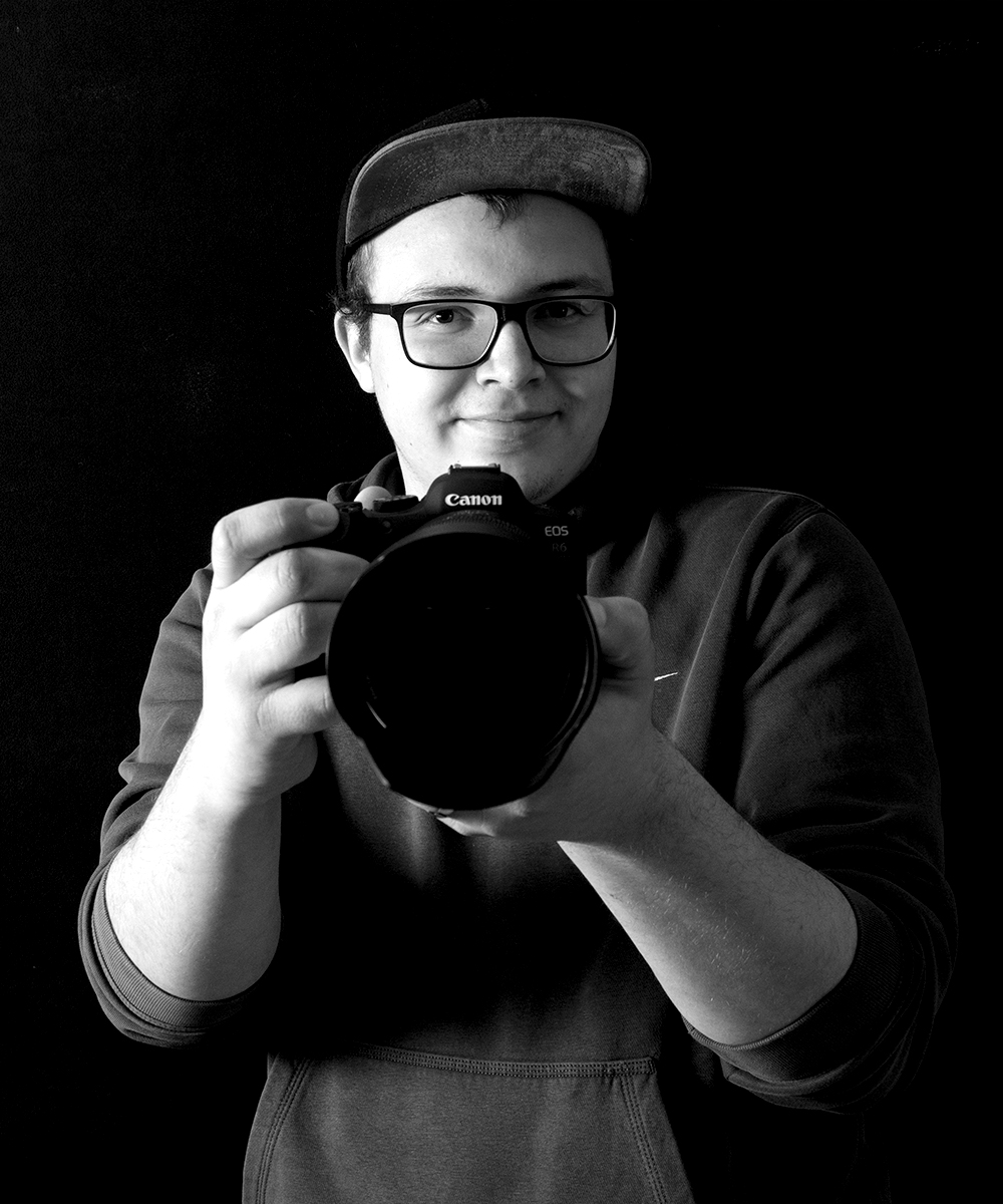 Schwarz-weiß Portrait von Teammitglied Nico Arndt, junger Mann mit Brille und Kappe, Blick in die Kamera, große Kamera in der Hand