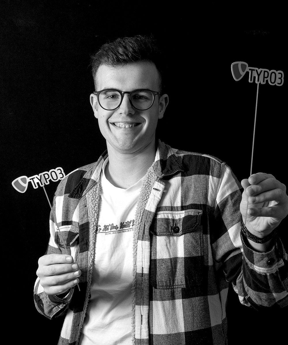 Schwarz-weiß Portrait von Teammitglied Luca Fink, junger Mann mit Brille und zwei Schildern in der Hand mit dem Schriftzug Typo3