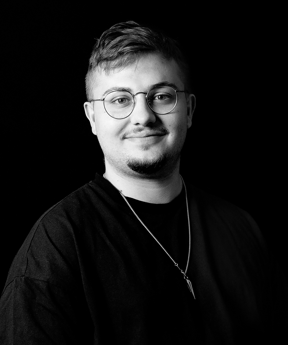 Schwarz-weiß Portrait von Teammitglied Jack Heidenwag, junger Mann mit Brille und Bart, Kette um den Hals