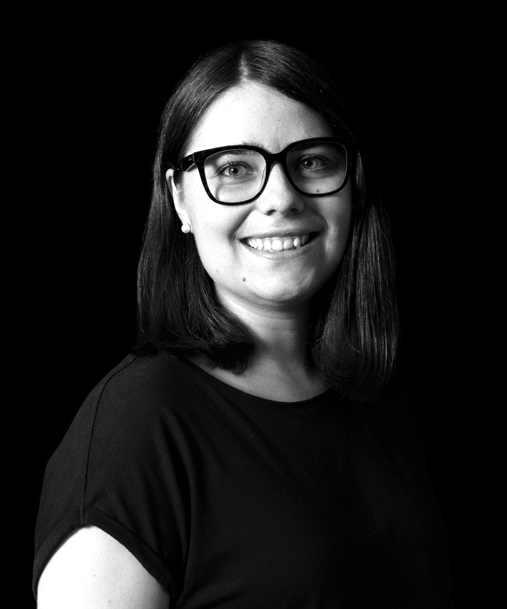 Schwarz-weiß Portrait von Teammitglied Sonja Mayer, junge Frau mit Ohrringen, Brille und kurzen Haaren, Blick in die Kamera